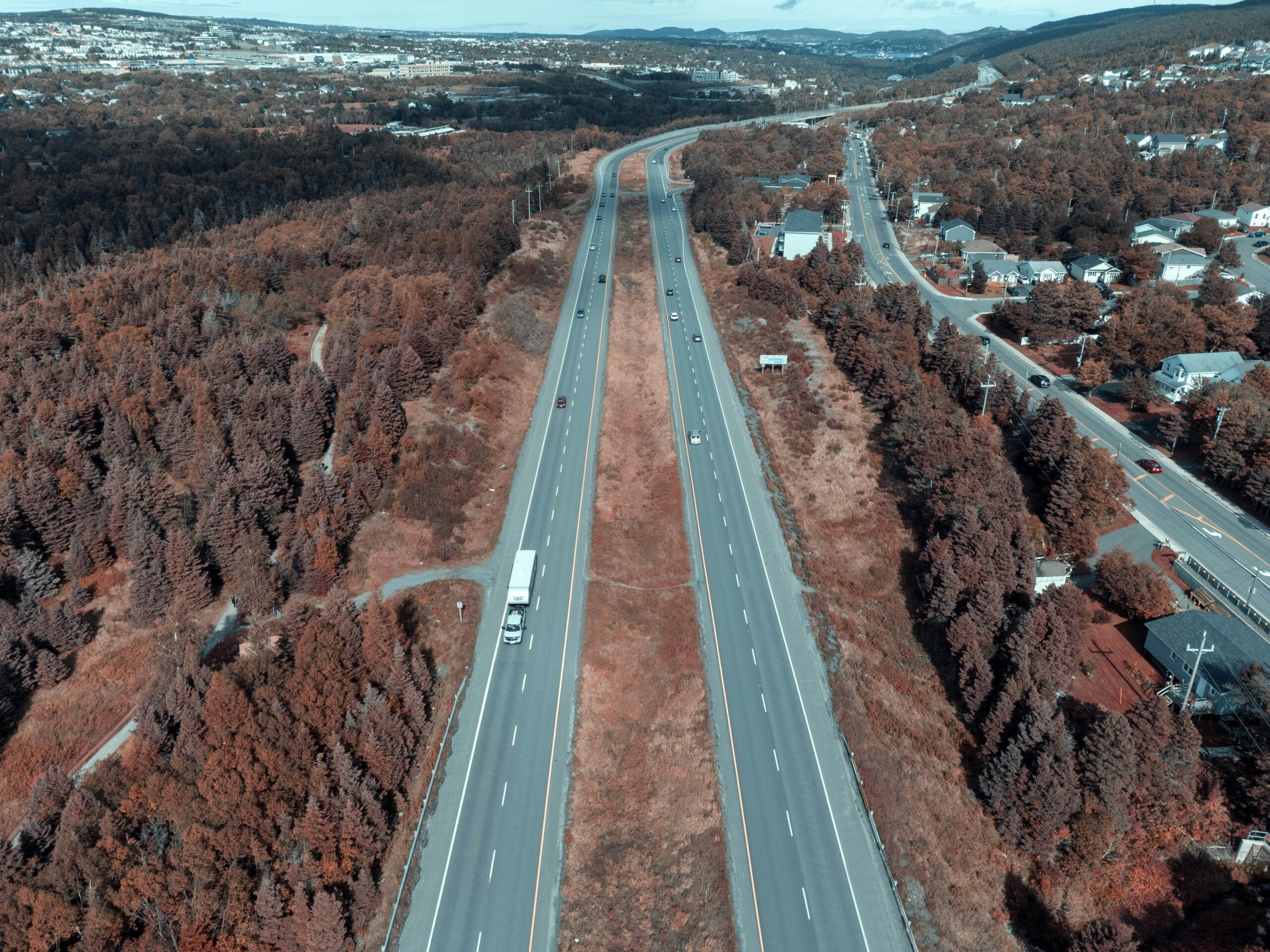 Aerial view of a divided highway running through a suburban area with houses on one side and forest on the other, showing traffic moving in both directions.