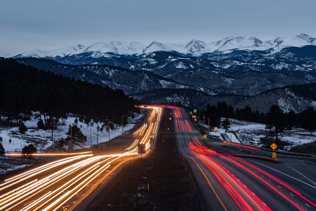 Long-exposure photo of highway traffic at night with light trails from cars leading into snow-covered mountains in the background.