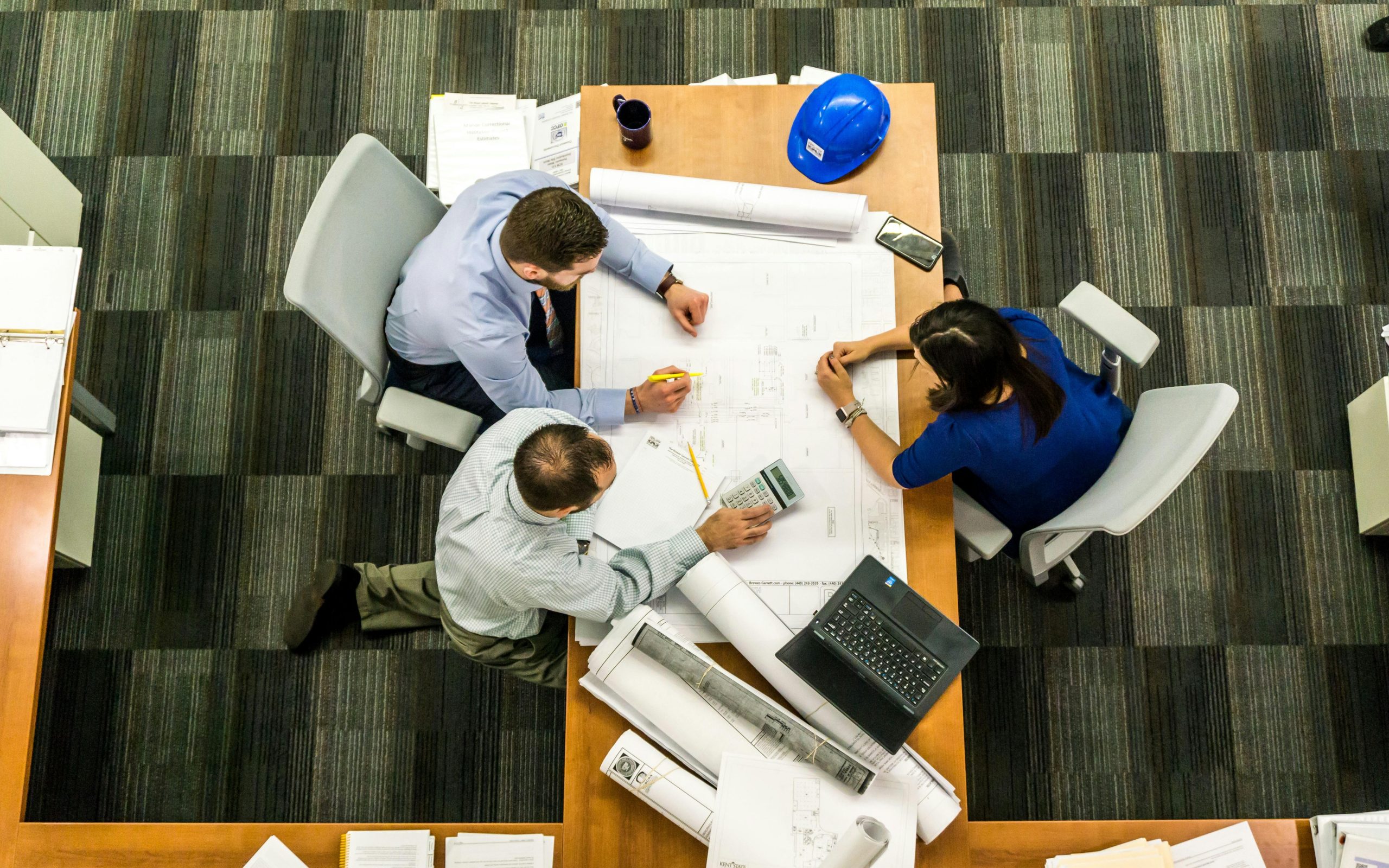 Aerial view of three construction managers sitting around a desk with blueprints and a calculator.