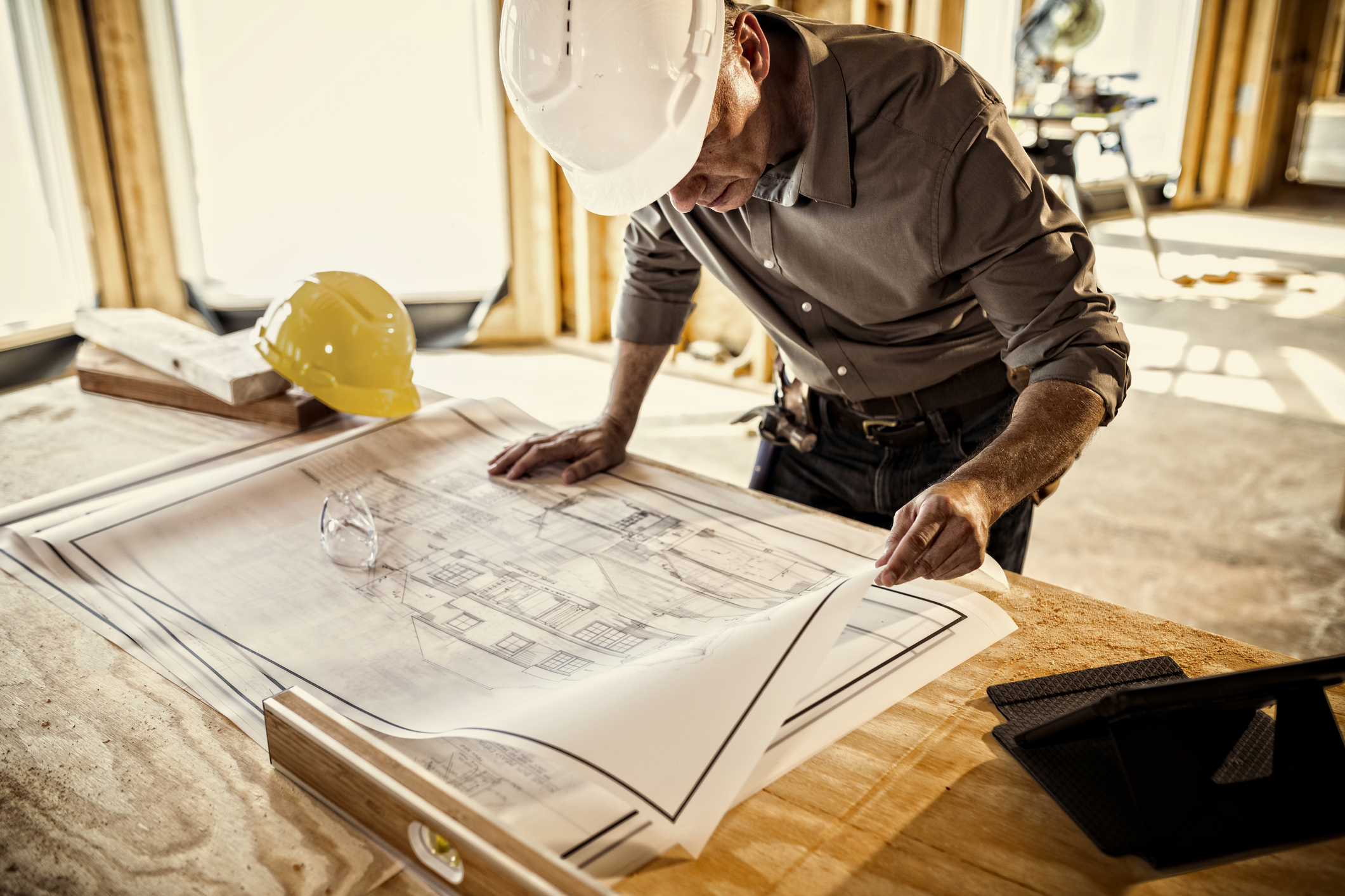 Builder in a white hard hat reviewing house plans inside a framed home, illustrating planning to build a stronger business in a tough market.
